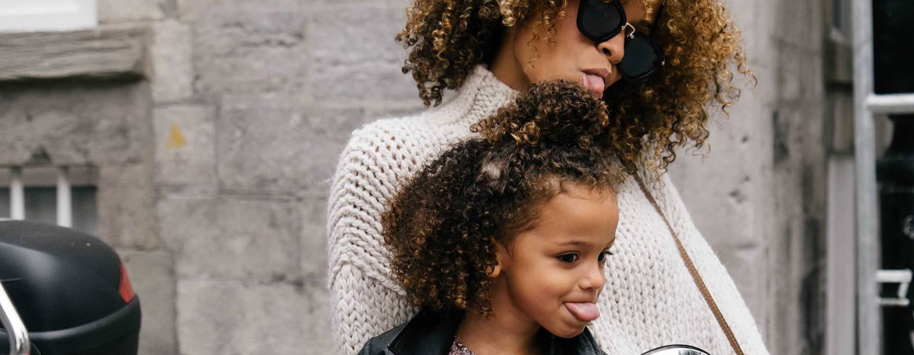 mother and daughter stick their tongues out while looking at themselves in a motorcycle mirror parked in front of a building